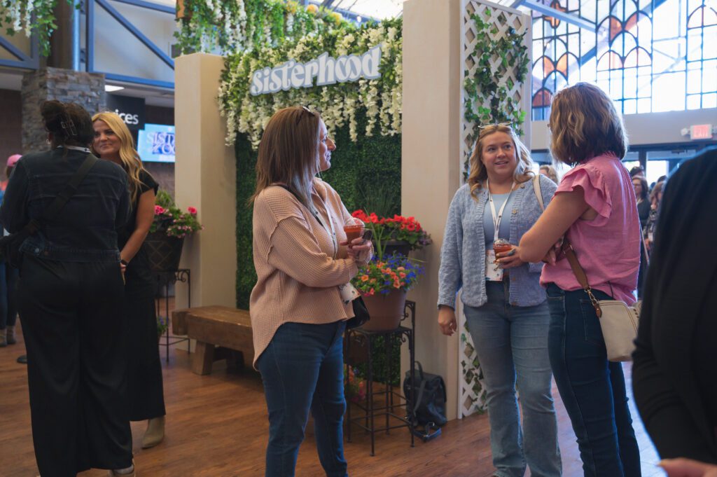 A group of women engage in conversation near a greenery wall with a “Sisterhood” sign overhead. Flower arrangements and hanging vines frame the space.