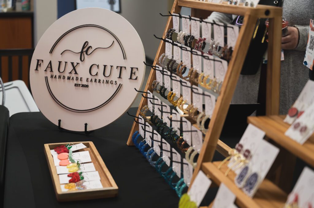 The Faux Cute vendor table with a wooden earring display rack, featuring glittery handmade hoop earrings and a branded circular sign.