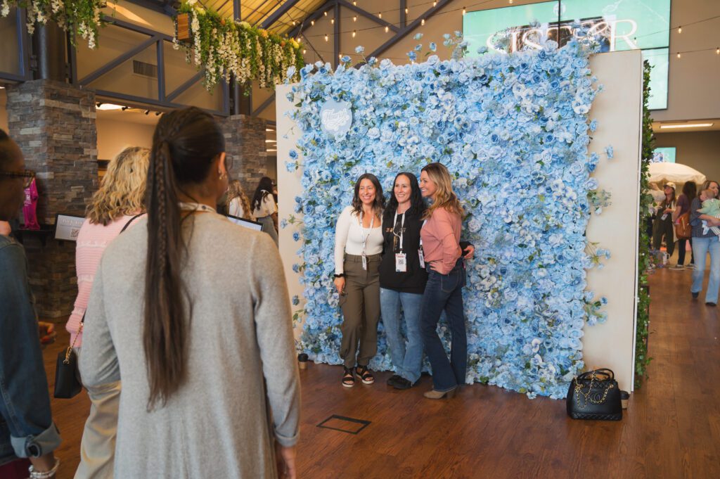 Women pose in front of a stunning blue flower wall at the Sisterhood event, surrounded by friends waiting their turn to take photos.