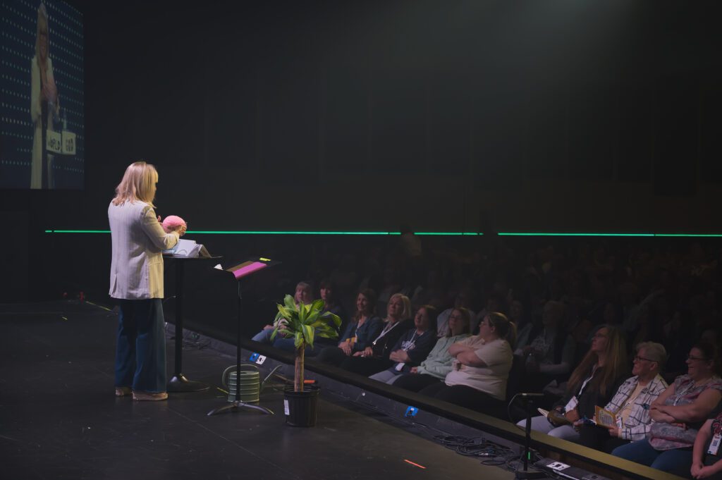 Ann Wilson on stage holding a brain prop while speaking to the audience at Sisterhood 2025, bringing humor and truth in a memorable moment.