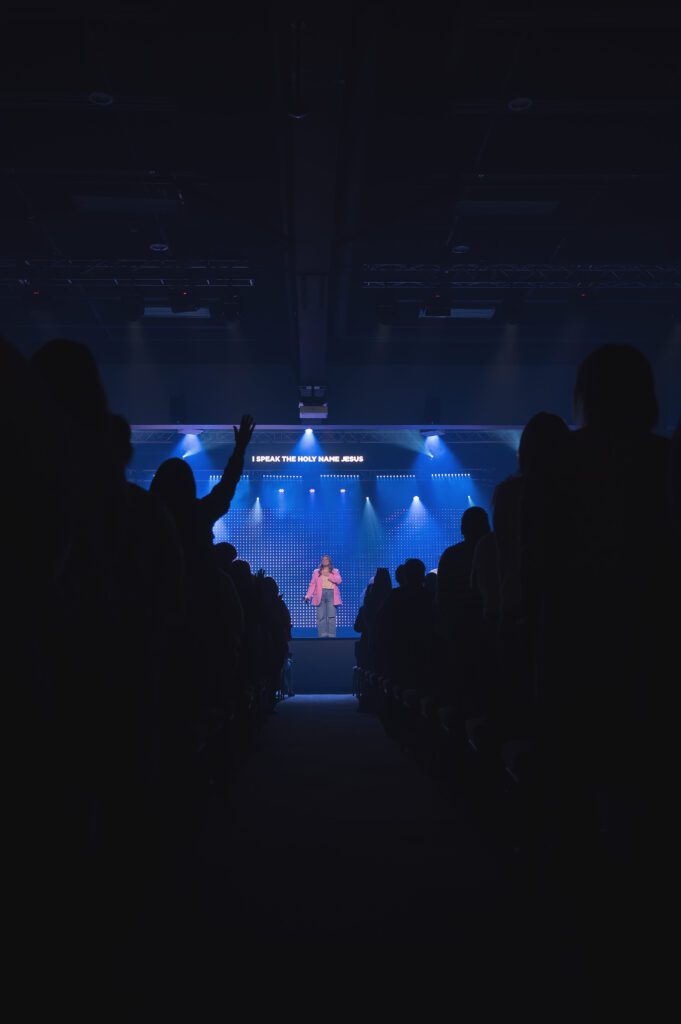 A worship singer leads the crowd in praise under bright blue lights with hands raised in the audience.