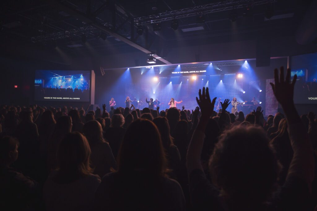 A full worship team leads the crowd in praise under bright lights with hands raised in the audience.