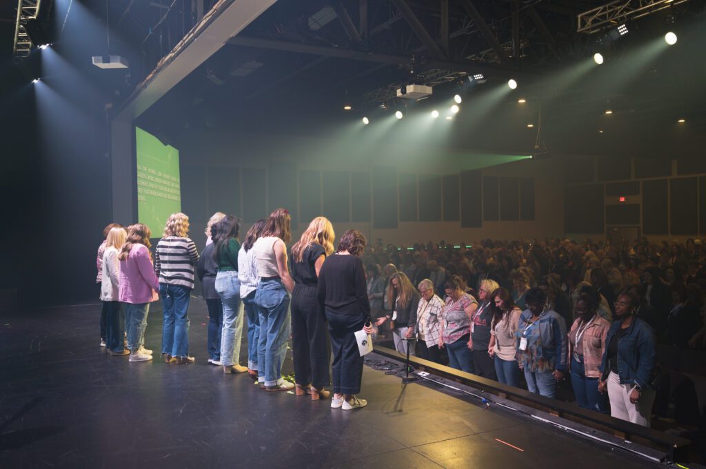 Women gather on stage in a powerful moment of prayer, heads bowed and arms wrapped around each other while the audience joins in silent support.